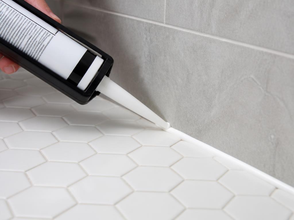 Caulking gun applying a clean white bead of caulk where gray wall tile meets white hexagon floor tile